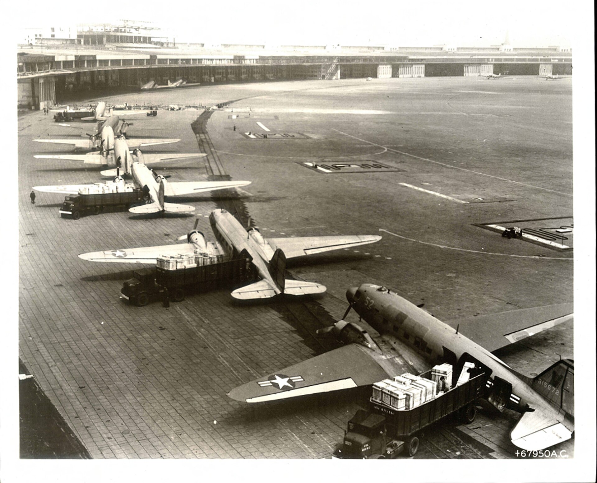 Several C-47 transport aircraft stand nose to tail while crews unload cargo across the Tempelhof airfield.