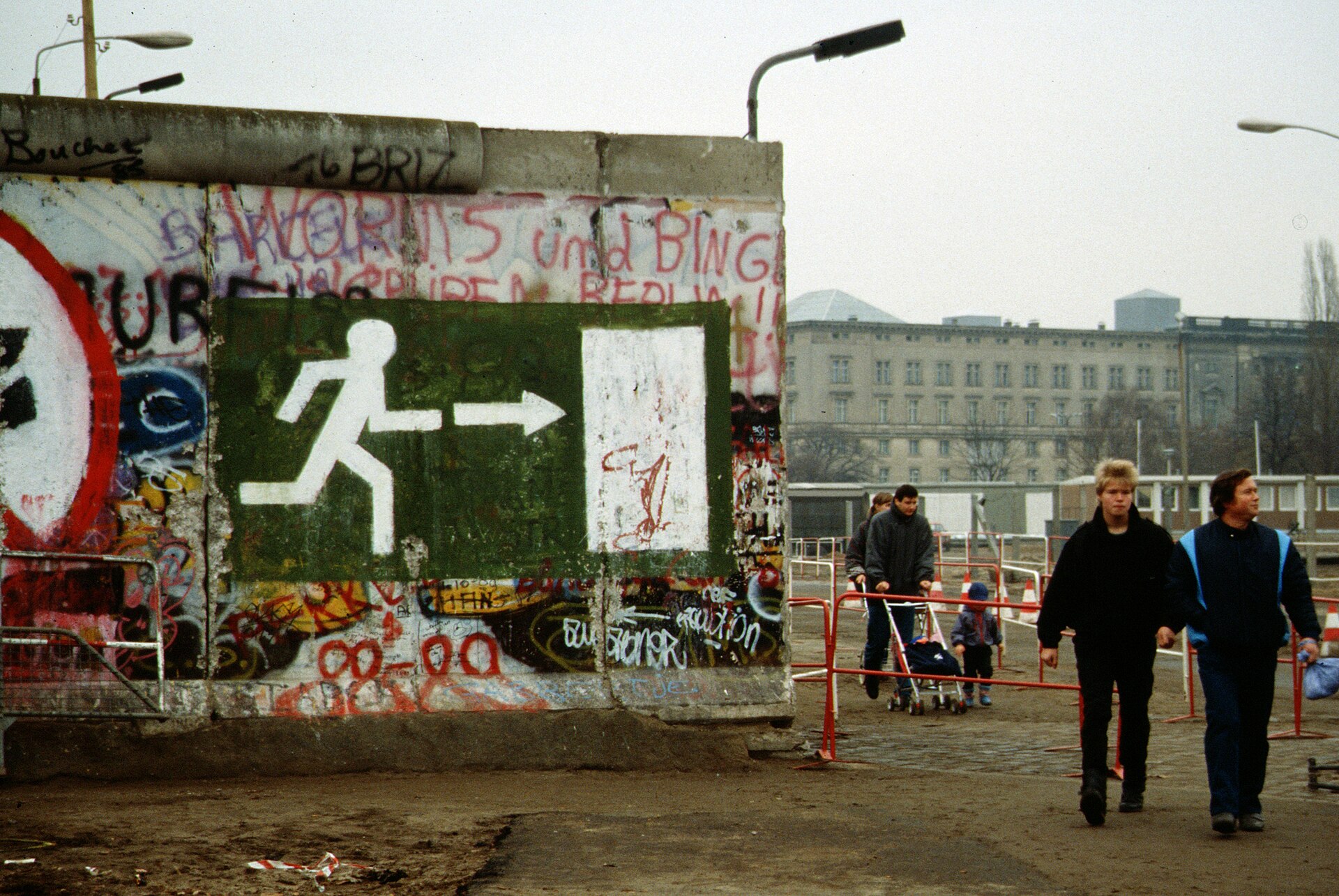 Pedestrians walk through an opening beside a graffiti covered wall panel marked with a white exit symbol.