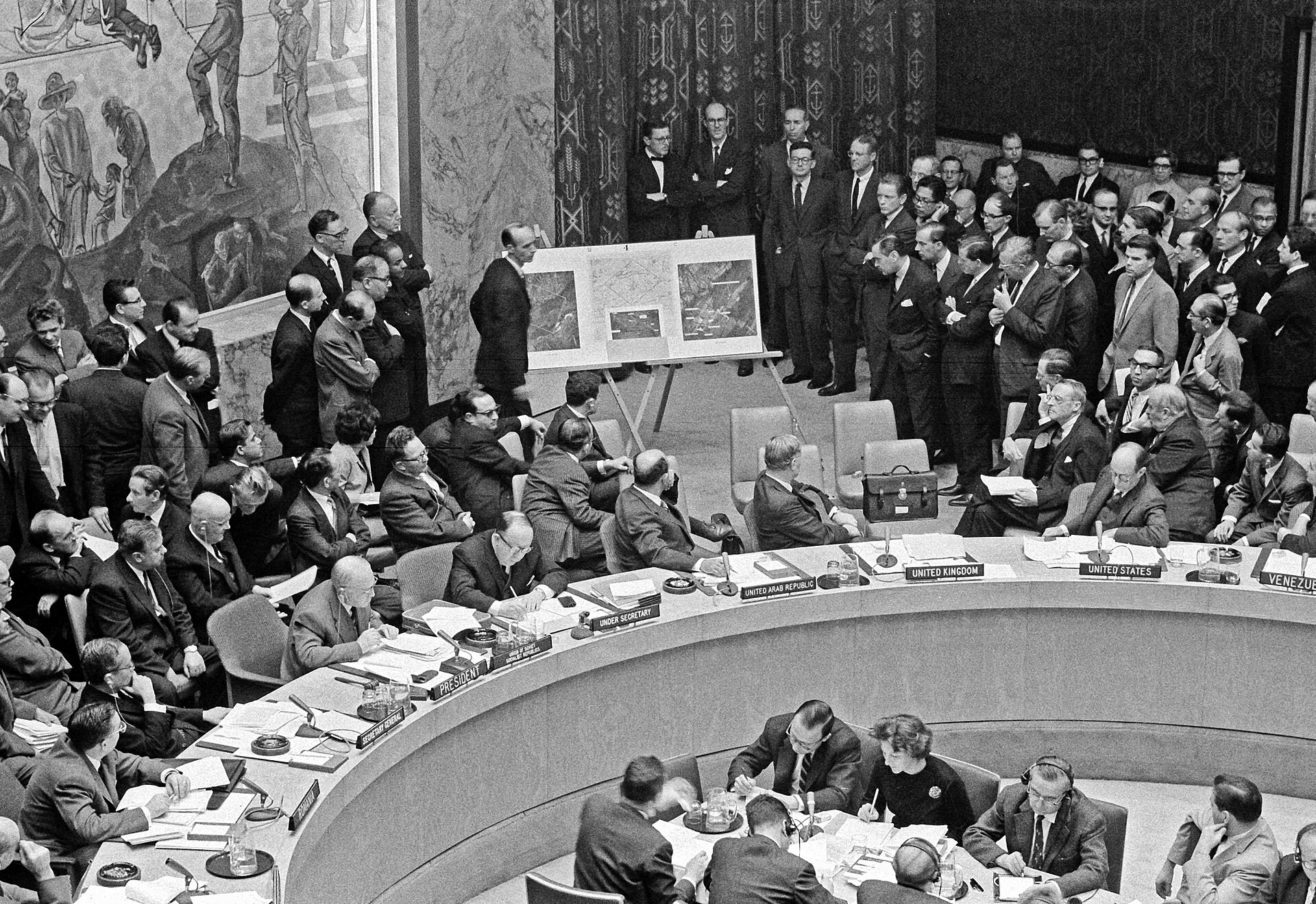 Adlai Stevenson points toward enlarged aerial missile photographs displayed before seated delegates in the UN Security Council chamber.