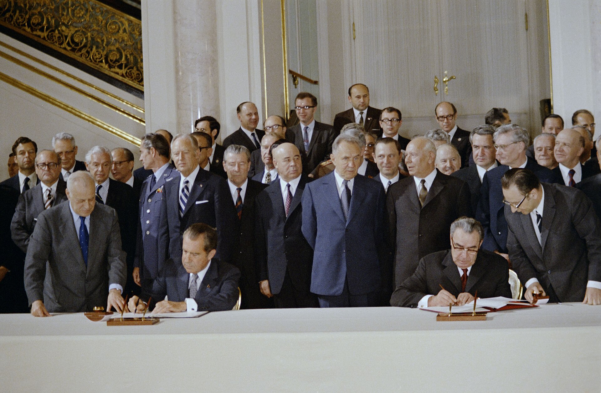 Richard Nixon and Leonid Brezhnev sit at a long table signing documents as officials stand behind them.
