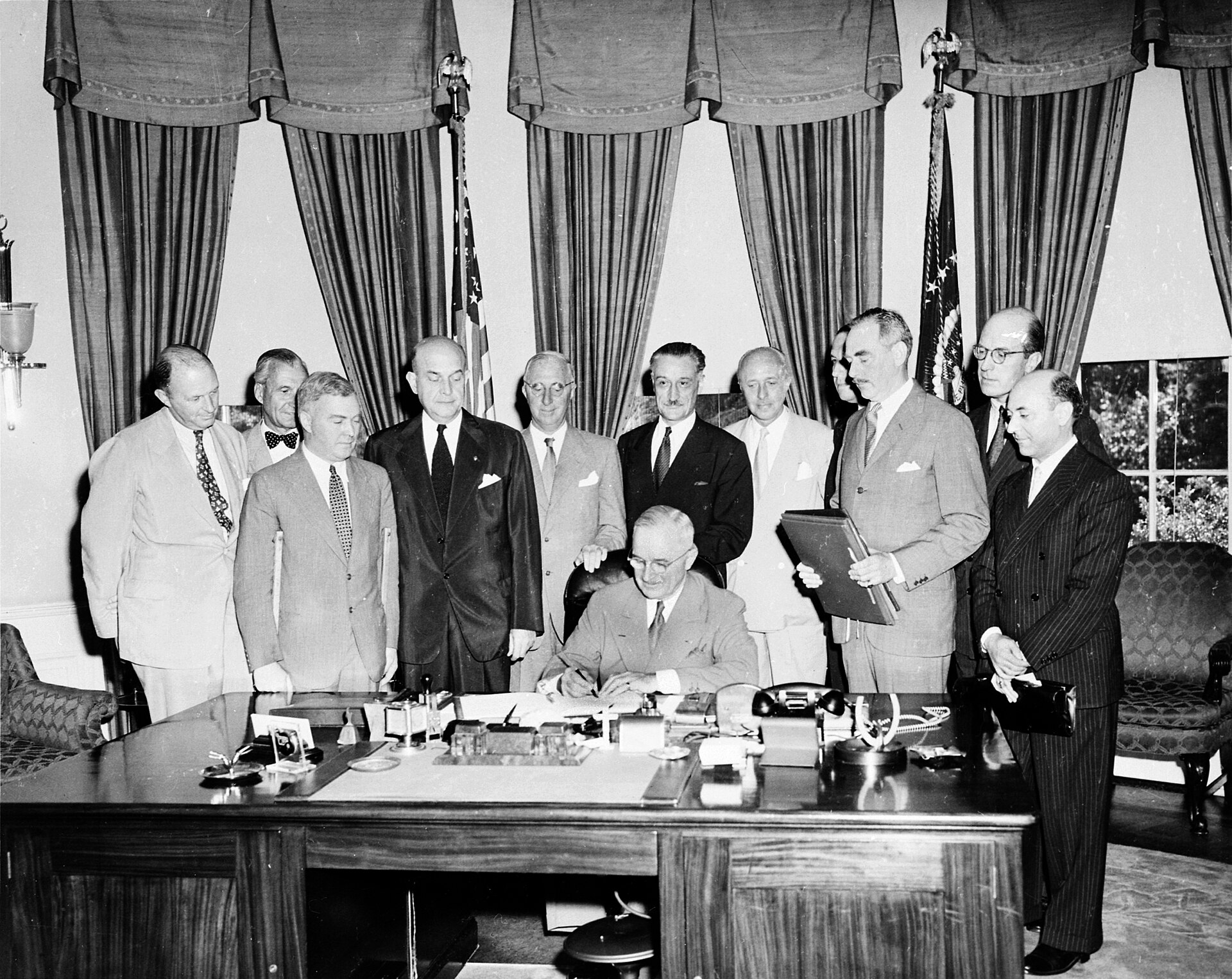 President Truman signs at his Oval Office desk while diplomats from several treaty states stand closely behind him.