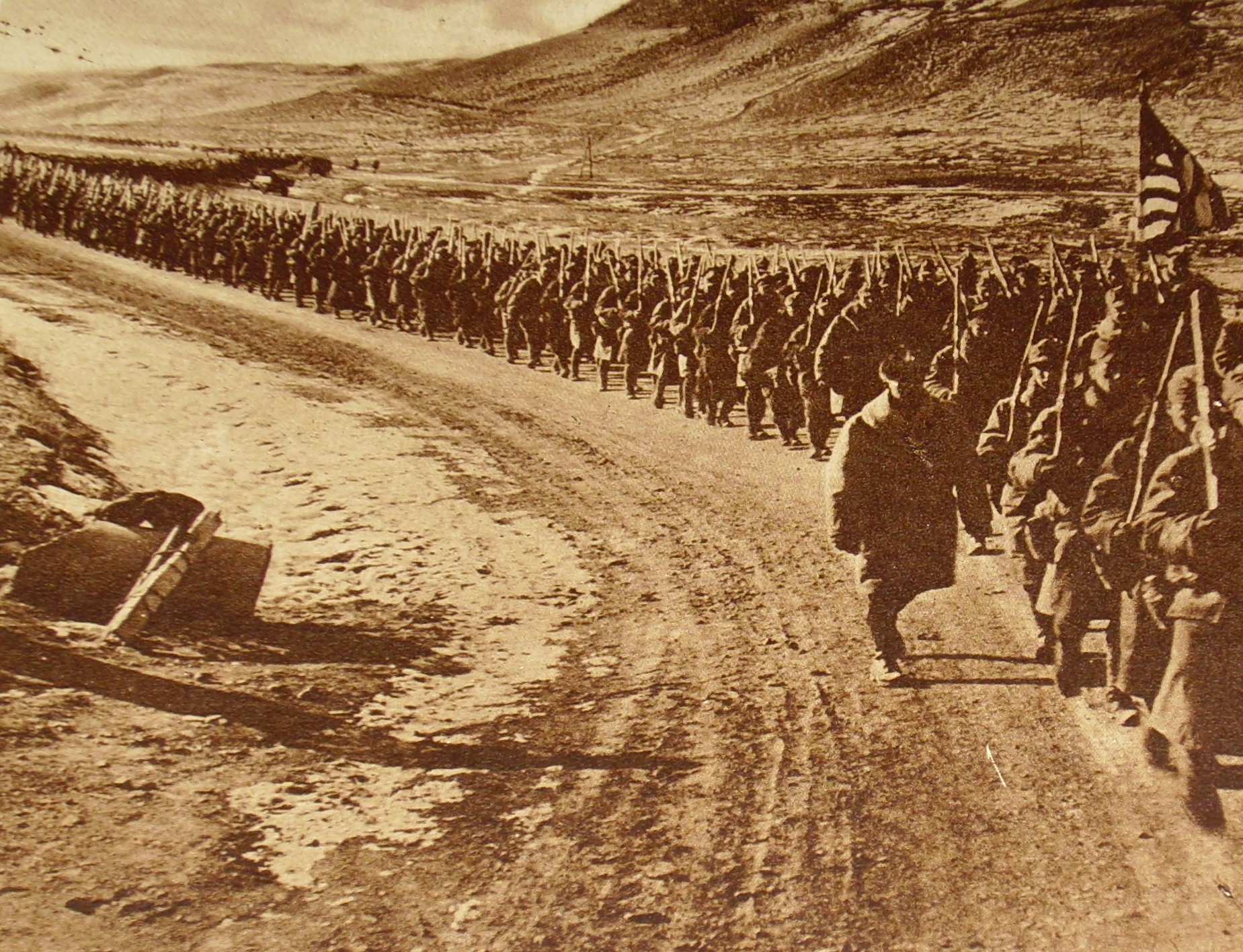 A long column of United States soldiers marches along a tree-lined road carrying packs and rifles.