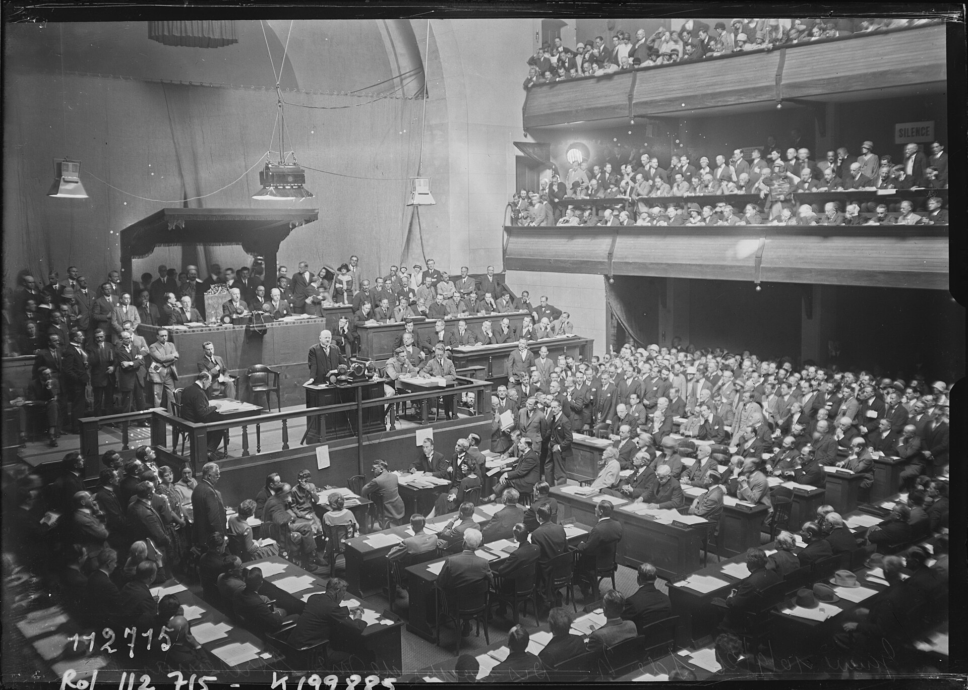 Gustav Stresemann speaks from a lectern to League delegates seated in a large hall.