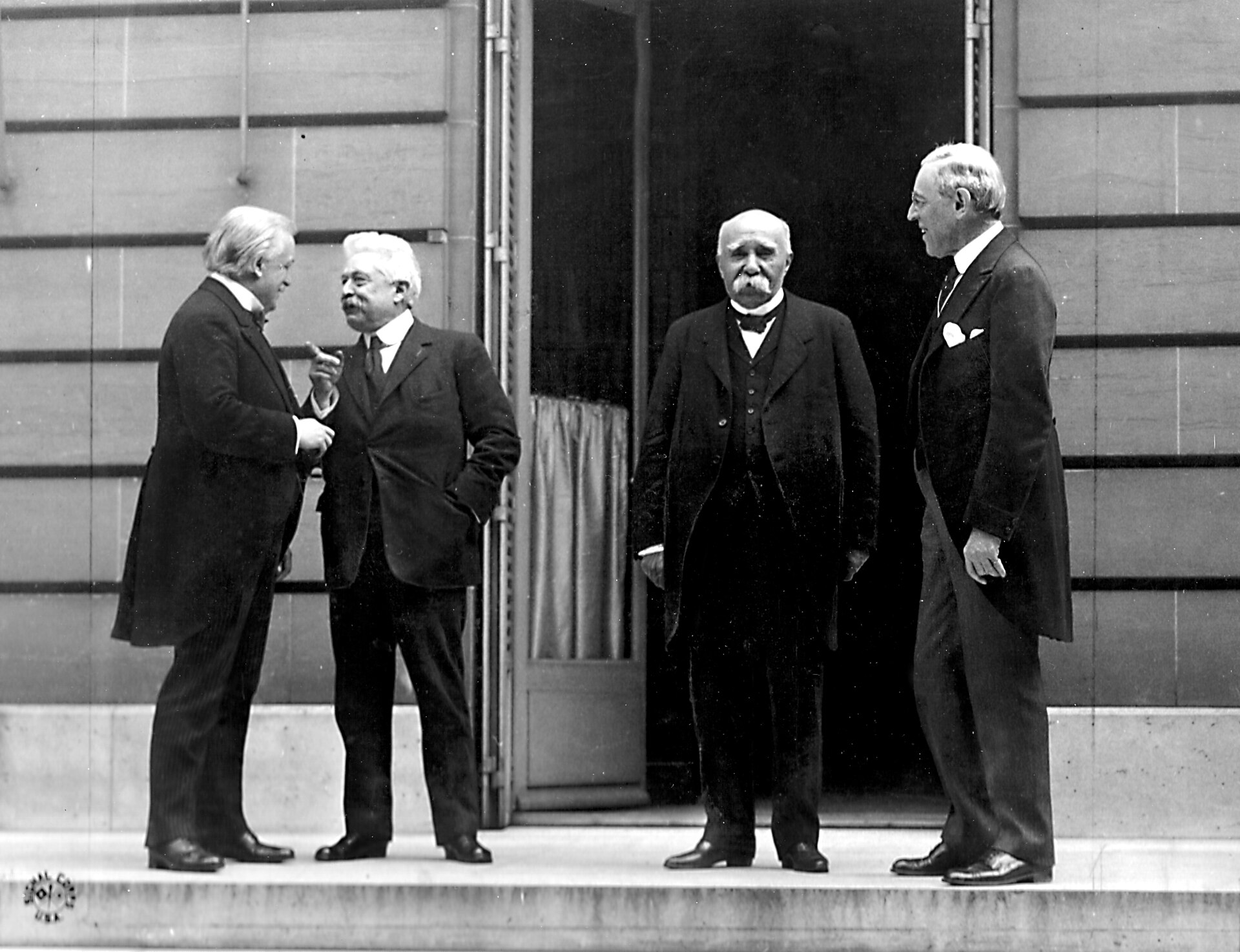 Lloyd George, Orlando, Clemenceau and Wilson stand together in dark suits for a photograph.