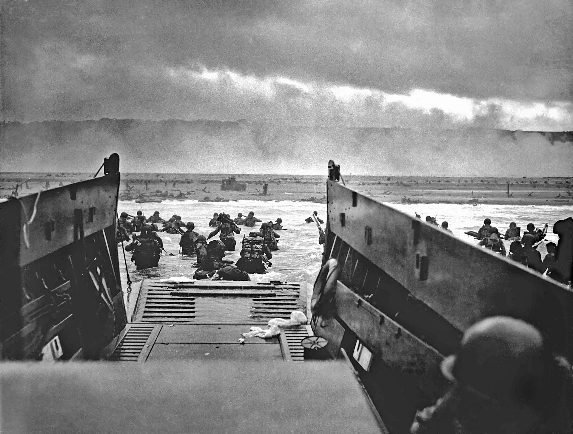 American soldiers wade through surf from a landing craft toward Omaha Beach under fire.