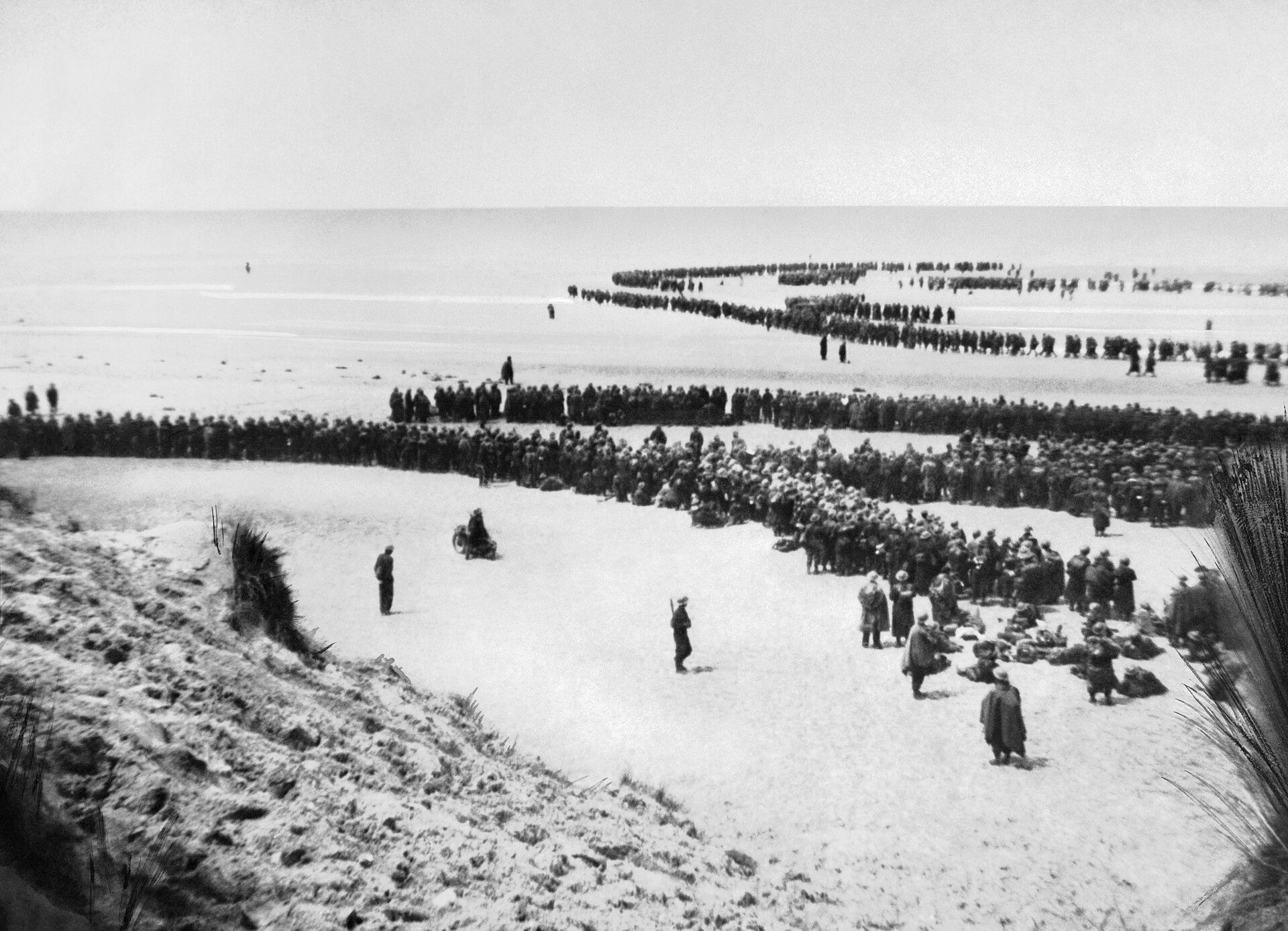 Long lines of British soldiers wait across the beach and shallow water at Dunkirk.