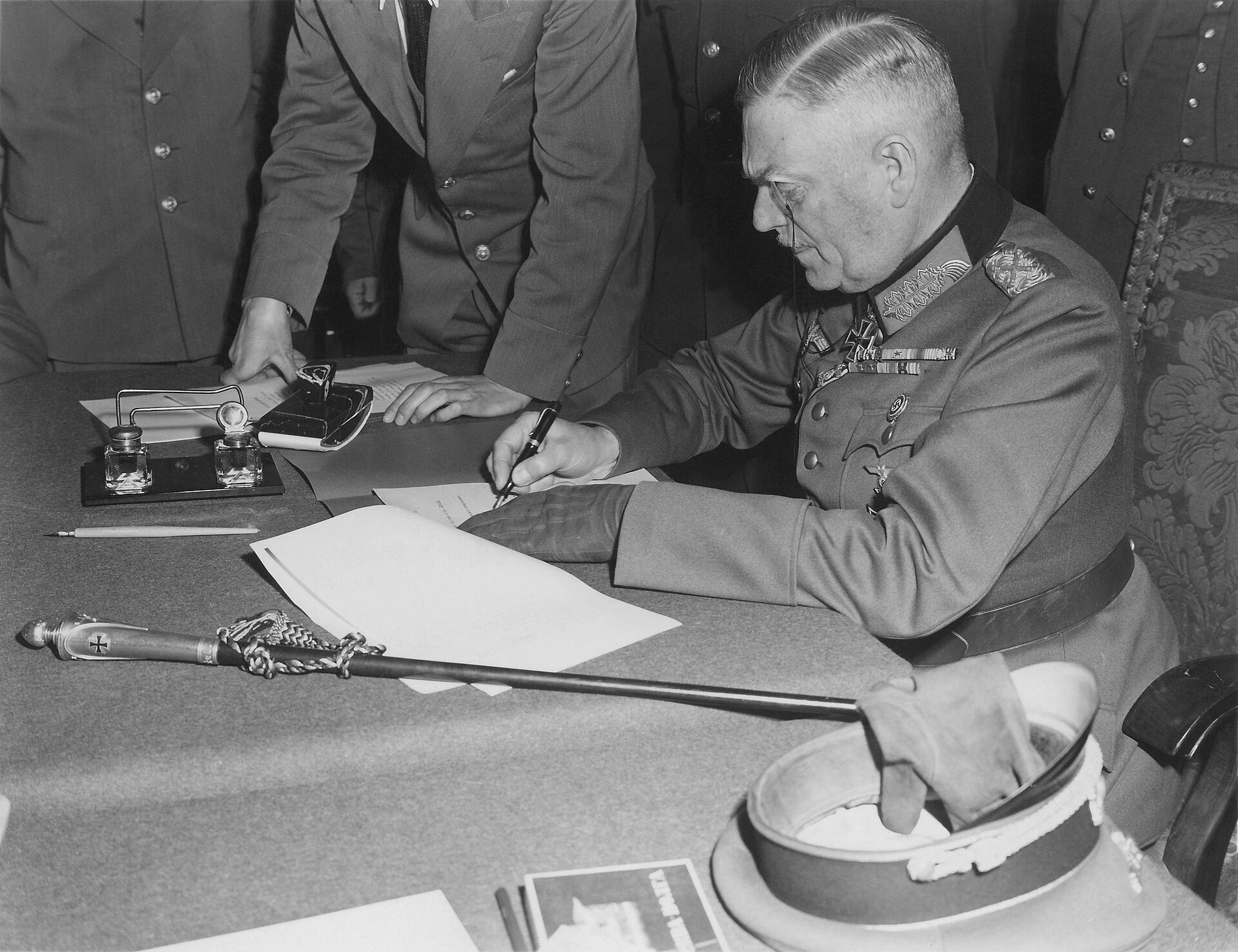 Field Marshal Wilhelm Keitel signs surrender documents at a table surrounded by Allied officers.